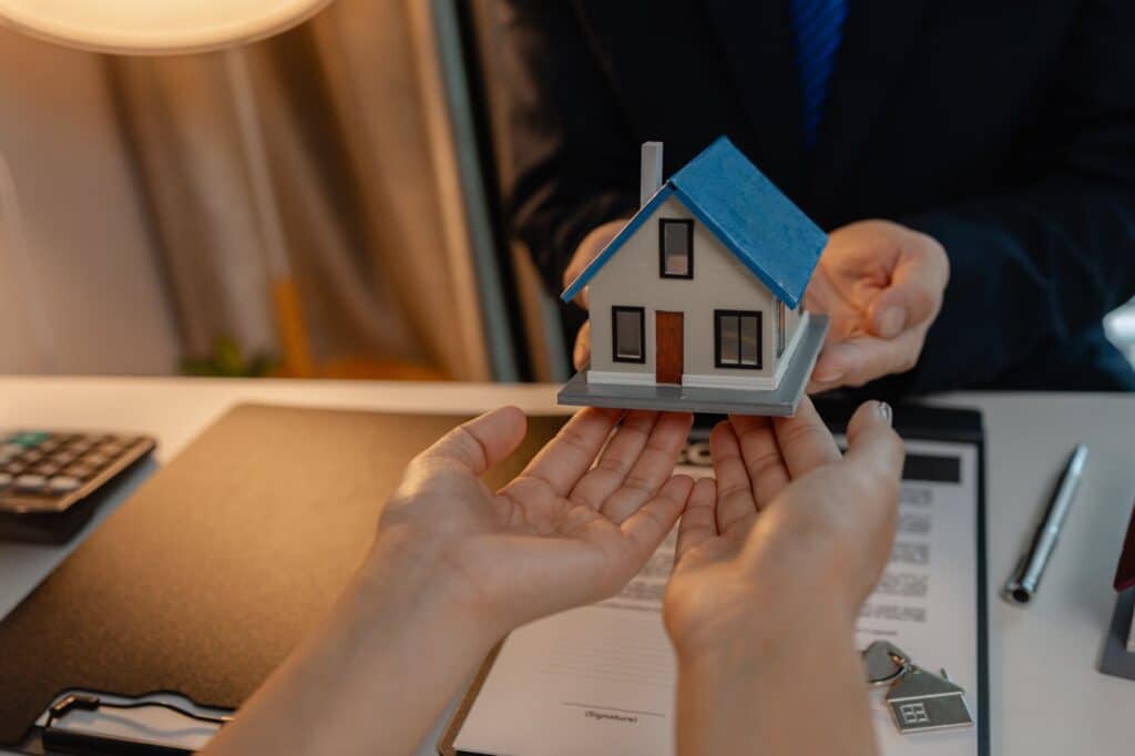 Person handing over a wooden house to another person across a desk with paperwork on it